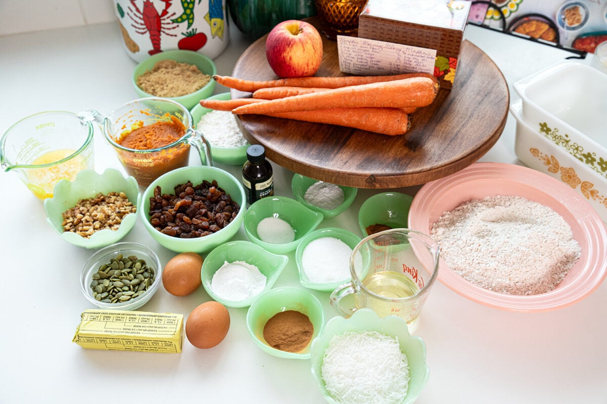 Various baking ingredients are arranged on a counter, including carrots, an apple, eggs, flour, sugar, spices, nuts, oil, raisins, coconut, and vanilla extract, set near measuring cups and a stick of butter.