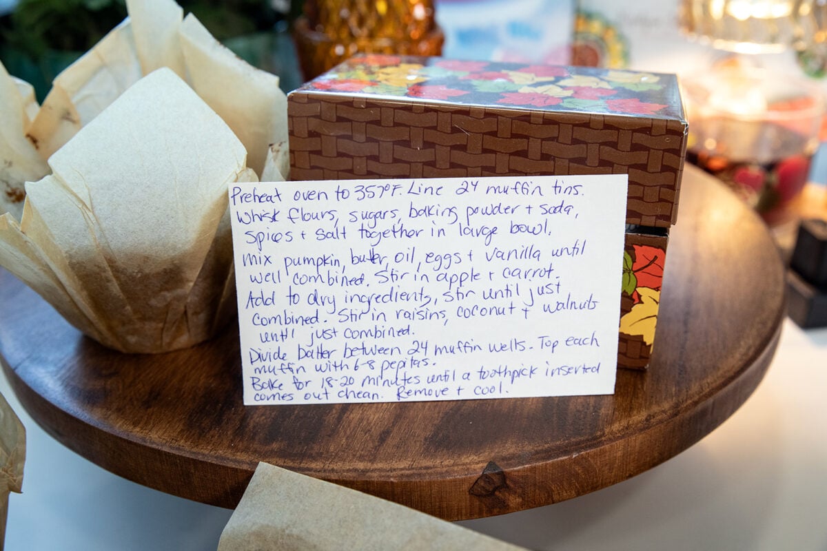 A handwritten recipe card for muffins sits on a wooden tray, surrounded by brown paper muffin liners and a decorative box with autumn leaves. The recipe details ingredients and baking instructions.