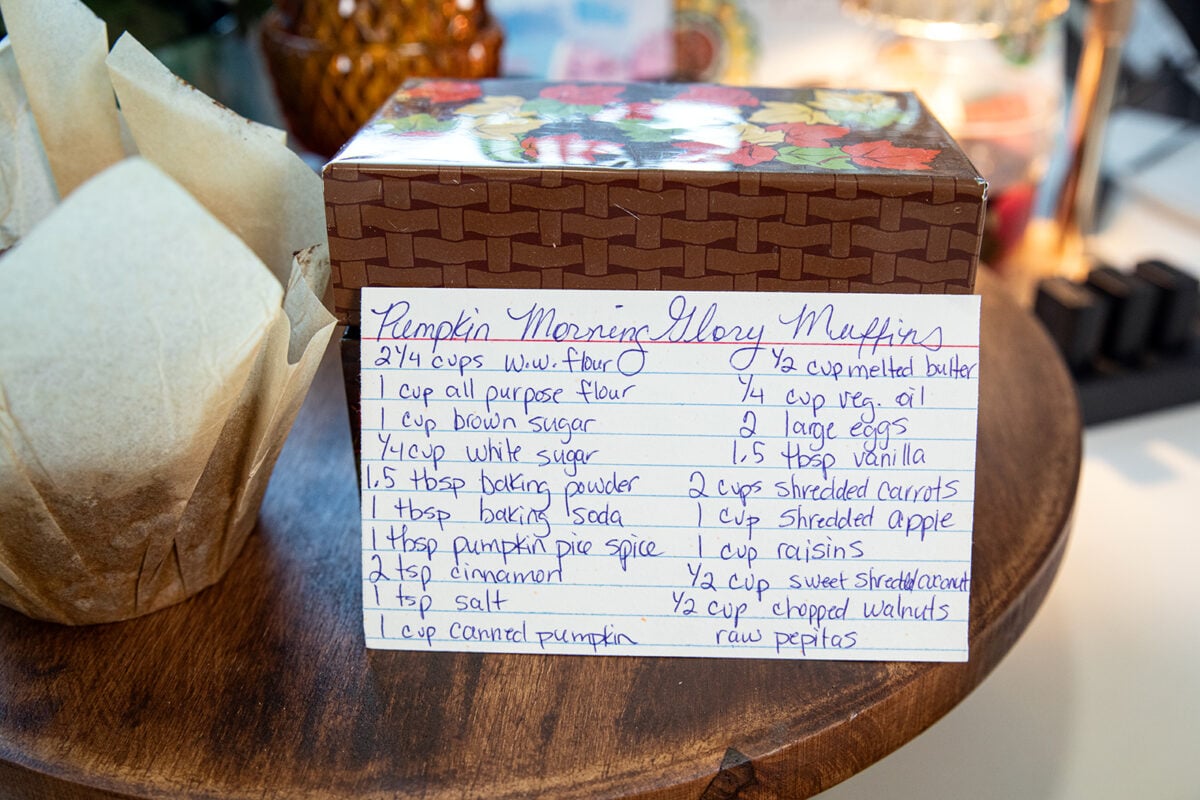 A handwritten recipe card for Pumpkin Morning Glory Muffins sits on a wooden table next to a paper towel holder. The recipe lists ingredients including flour, sugar, eggs, spices, pumpkin, apples, coconut, and walnuts.