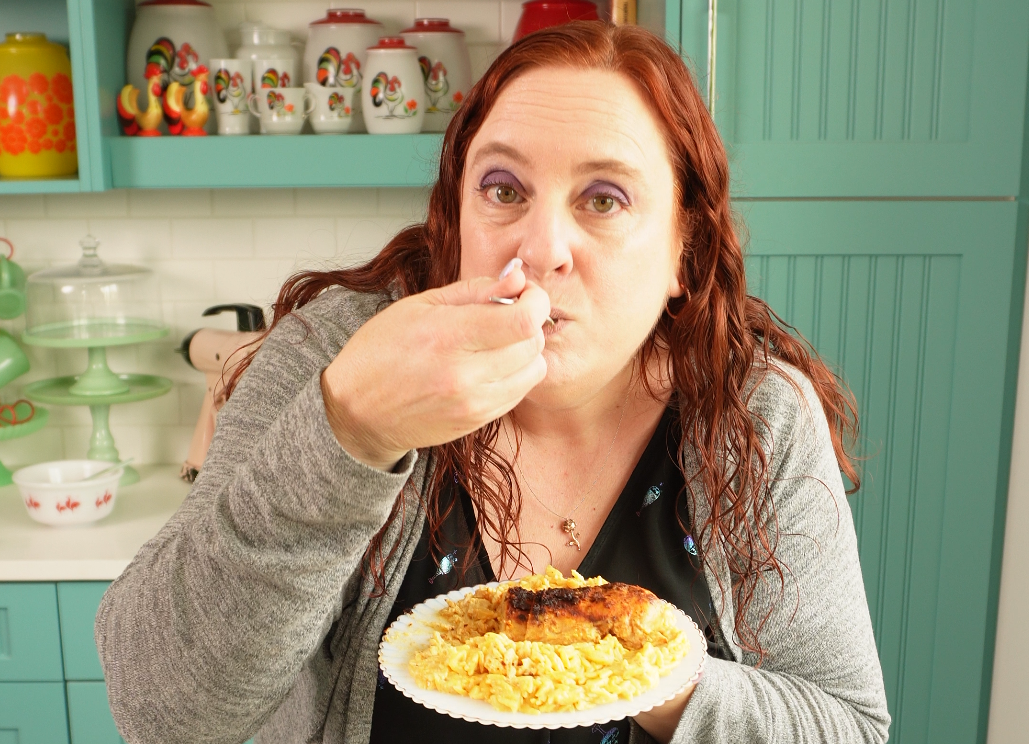 A woman with long red hair eats from a plate of rice and chicken in a colorful kitchen with teal cabinets and fruit-themed jars in the background.