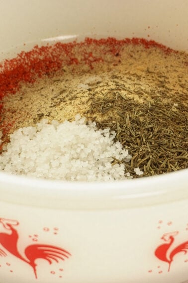 A white bowl with red rooster designs holds small piles of various spices, including blackened chicken seasoning, red powder, dried herbs, ground pepper, and coarse salt.