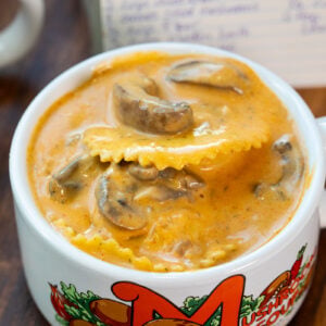 A bowl of creamy mushroom soup with ravioli, served in a white mug decorated with colorful mushrooms and the word Mushroom. A handwritten recipe card is visible in the background.