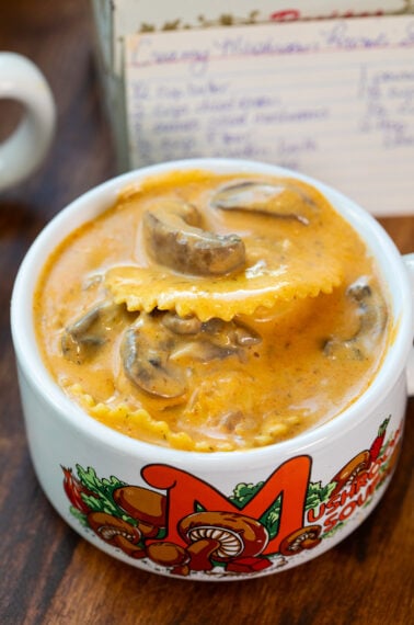 A bowl of creamy mushroom soup with ravioli, served in a white mug decorated with colorful mushrooms and the word Mushroom. A handwritten recipe card is visible in the background.