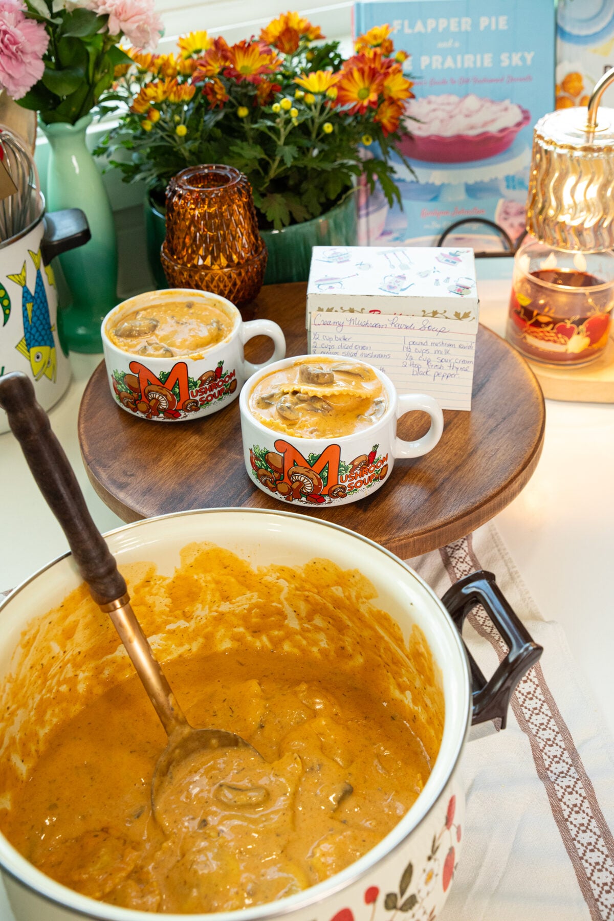 Two mugs filled with creamy soup sit on a wooden tray next to a pot of the same soup, flowers, and a cookbook. The setting feels cozy and inviting, with warm lighting and kitchen decor.