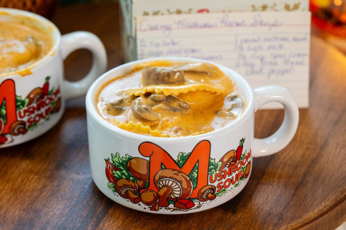 A white cup labeled Mushroom Soup is filled with creamy mushroom ravioli soup. Another similar cup and a handwritten recipe card are in the background on a wooden surface.