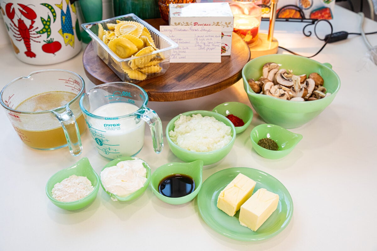 Various ingredients for a recipe are arranged on a kitchen counter, including pasta, chopped onions, mushrooms, butter, cream, flour, broth, sour cream, soy sauce, and spices in green glass bowls and measuring cups.