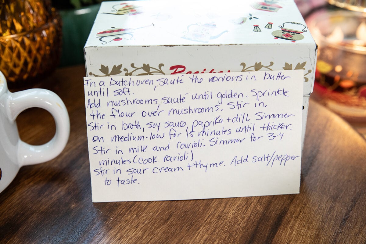 A handwritten recipe card on a white box, detailing steps to cook beef with mushrooms, broth, soy sauce, paprika, ravioli, and sour cream, with a mug and festive decor in the background.