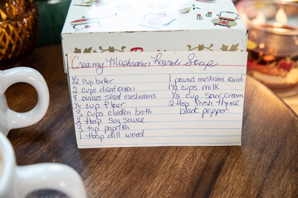 A handwritten recipe card for creamy mushroom ravioli soup is propped against a festive box, with cups and table décor visible beside it. The card lists ingredients such as butter, onion, mushrooms, and spices.