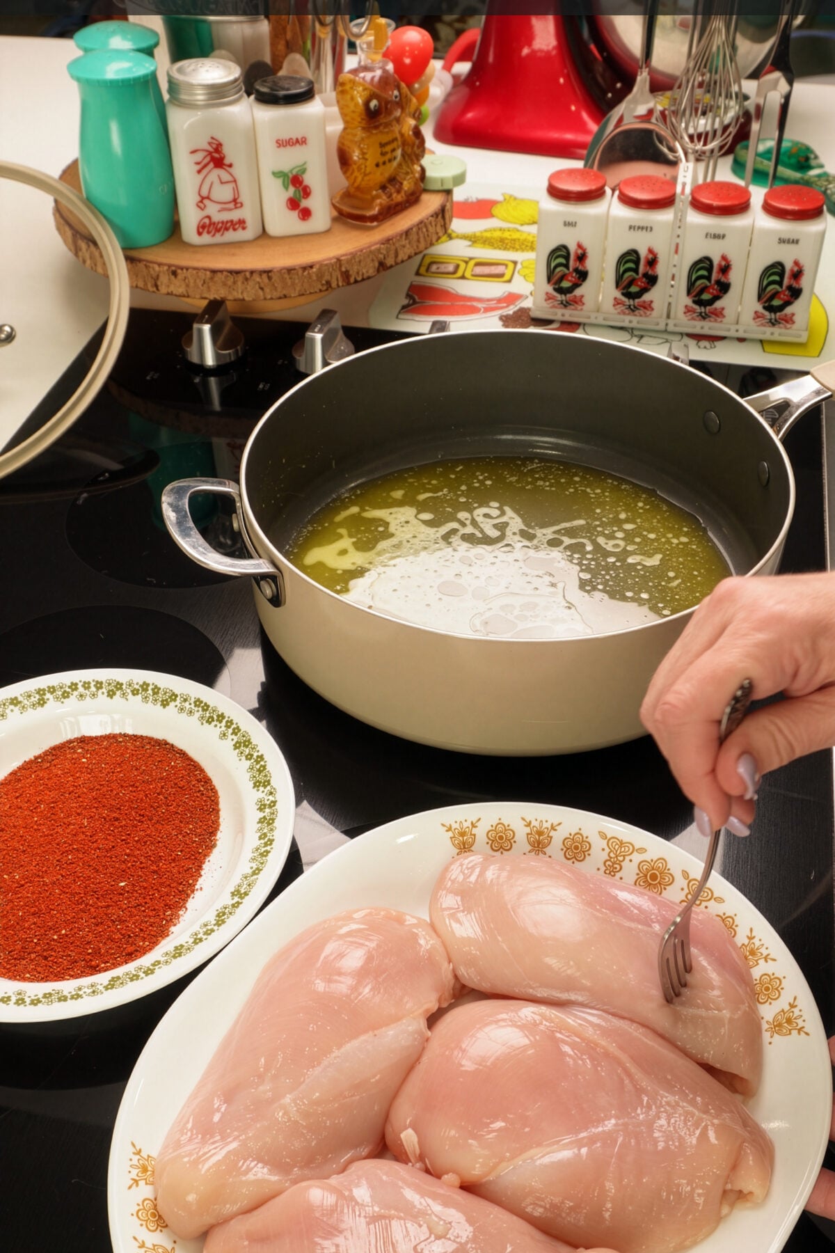 A hand pierces raw chicken breasts with a fork next to a bowl of red blackened chicken seasoning and a pan of oil on the stove, surrounded by spice jars and kitchen tools.