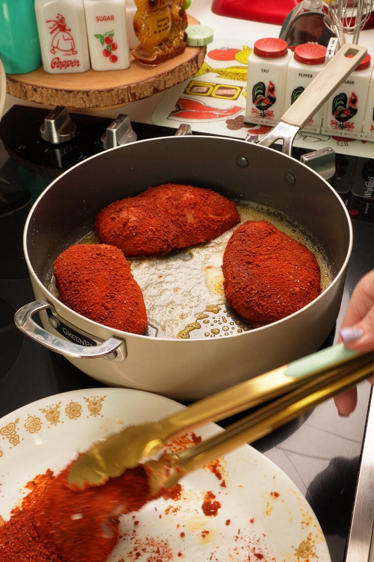 Three breaded chicken breasts cook in a pan with bubbling oil on a stovetop while a hand uses tongs to add another piece from a plate. Various spice containers, including blackened chicken seasoning, are visible in the background.