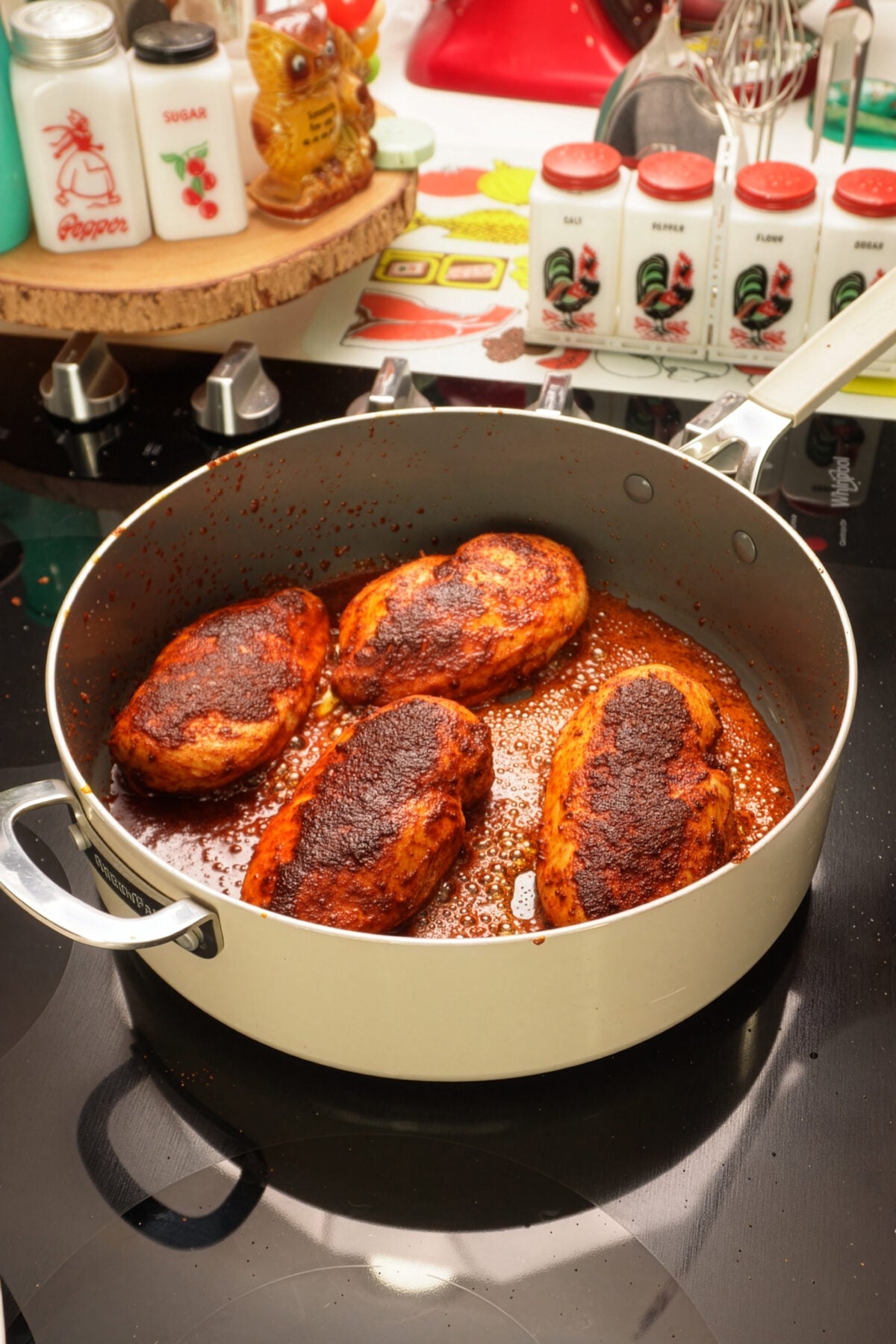 Four seasoned chicken breasts are cooking in a beige skillet on a stovetop, generously coated in blackened chicken seasoning. Colorful vintage spice containers and kitchen decor are visible in the background.