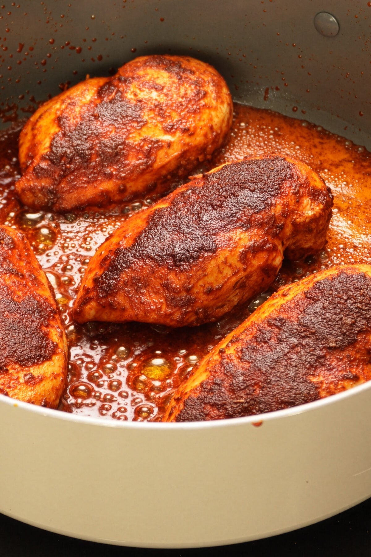 Four seasoned chicken breasts, coated in a rich blackened chicken seasoning, are cooking in a pan with bubbling sauce. The chicken is enveloped in a dark, reddish-brown spice rub, while the flavorful sauce pools around the pieces.