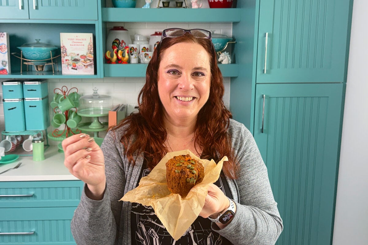 A woman with long red hair smiles while holding a large muffin in parchment paper in a retro-style kitchen with teal cabinets and colorful decor.