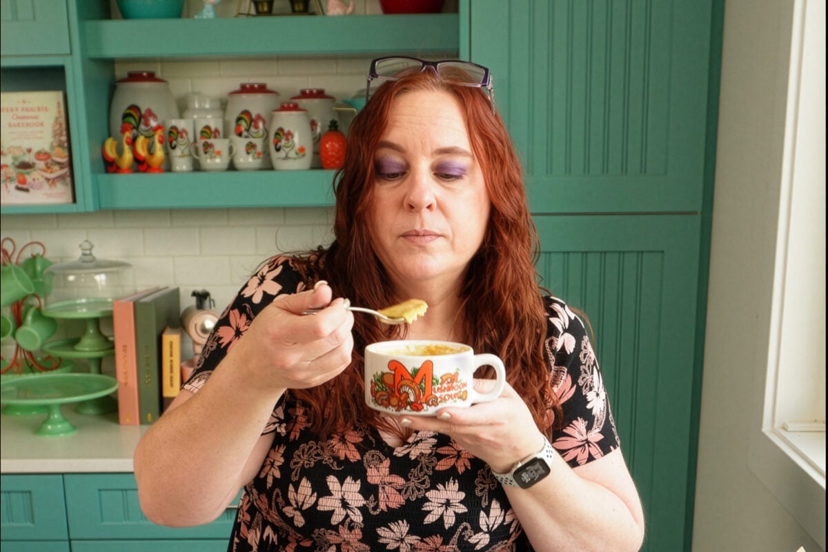 A woman with long red hair eats soup from a floral mug in a colorful kitchen with teal cabinets and rooster-themed decor.