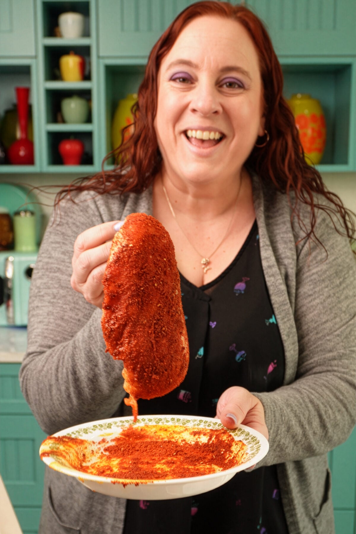 A smiling woman in a kitchen holds up a piece of seasoned meat over a plate, coating it with vibrant blackened chicken seasoning and bright red spices. Colorful dishes are visible on teal shelves in the background.