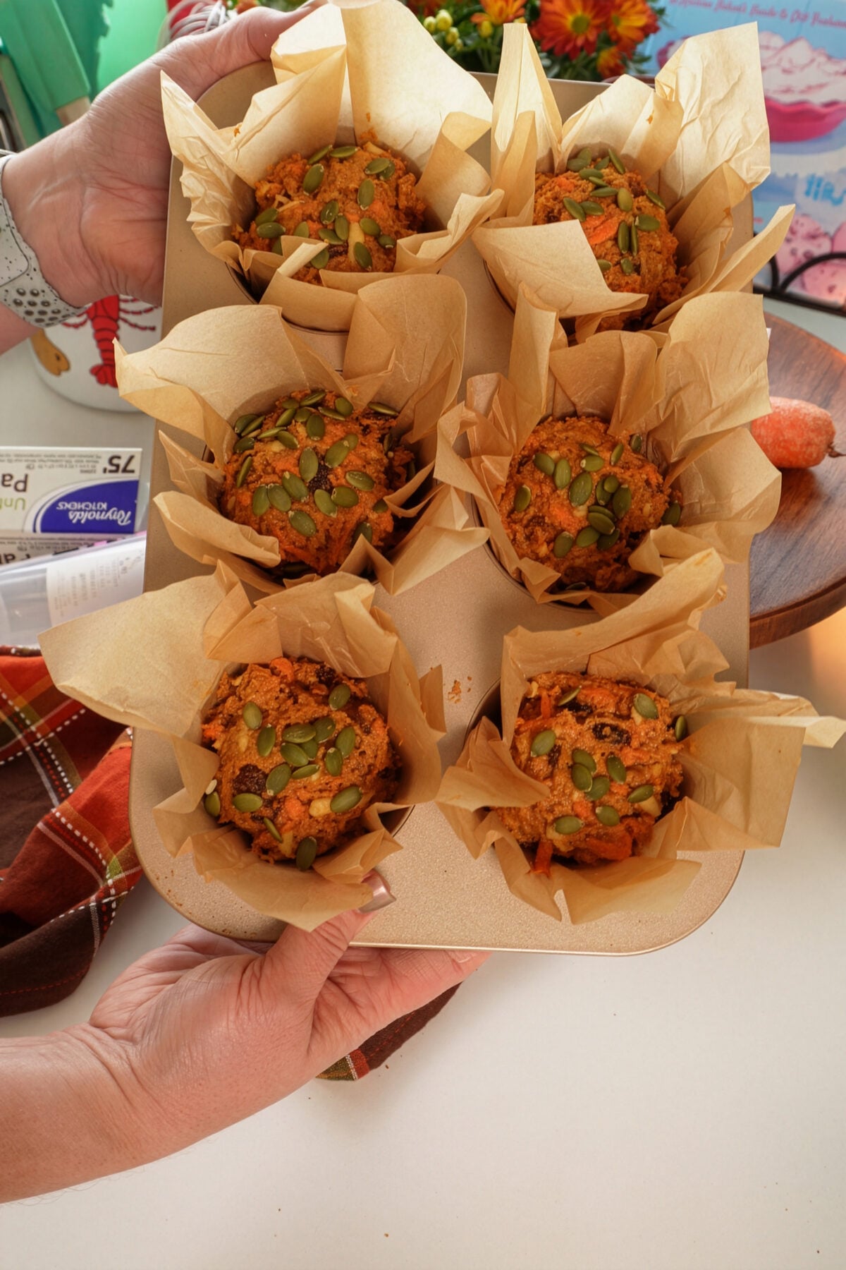 A person holds a muffin tin filled with six muffins in brown parchment paper liners, topped with pumpkin seeds and crumbs.