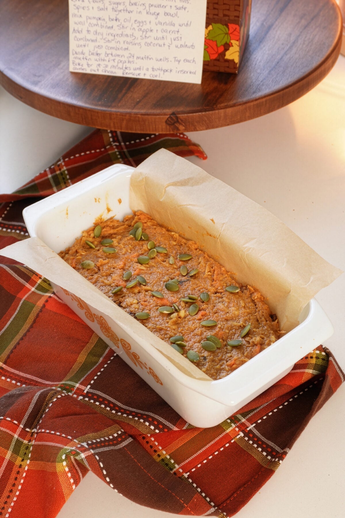 A loaf of pumpkin bread topped with pumpkin seeds sits in a parchment-lined baking dish on a red plaid cloth, with a handwritten recipe card in the background.