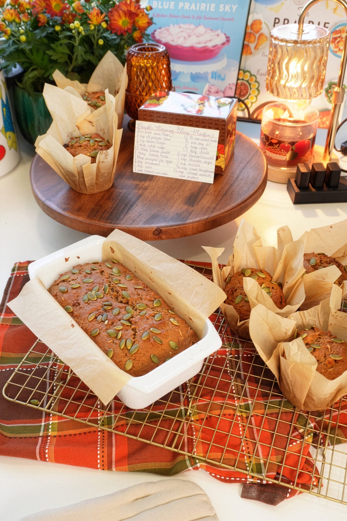 A loaf of pumpkin bread in a white pan and four muffins with pumpkin seeds rest on a cooling rack, surrounded by autumn decor, flowers, a recipe card, and a colorful cloth.