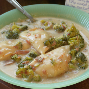 A green plate of creamy chicken and broccoli, garnished with small bacon pieces, with a spoon resting in the dish. Handwritten recipe notes are visible in the background.