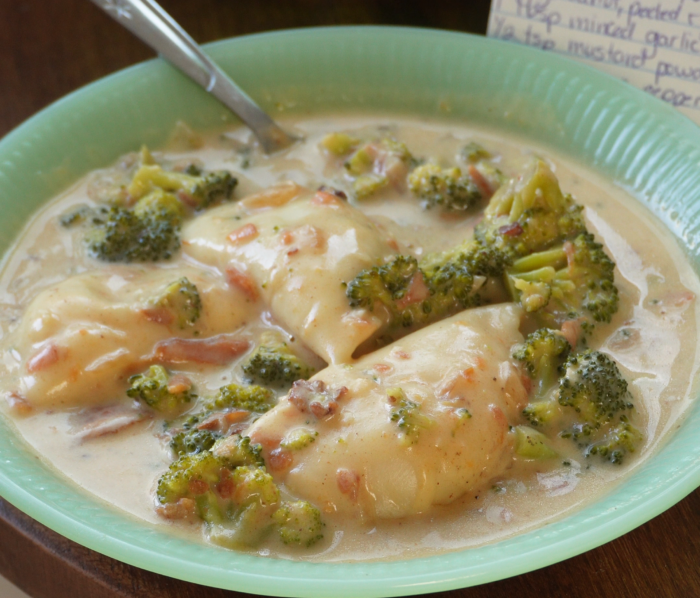 A green plate filled with creamy chicken breasts, broccoli, and bits of bacon in a white sauce. A fork rests on the plate. A handwritten recipe is partially visible in the background.