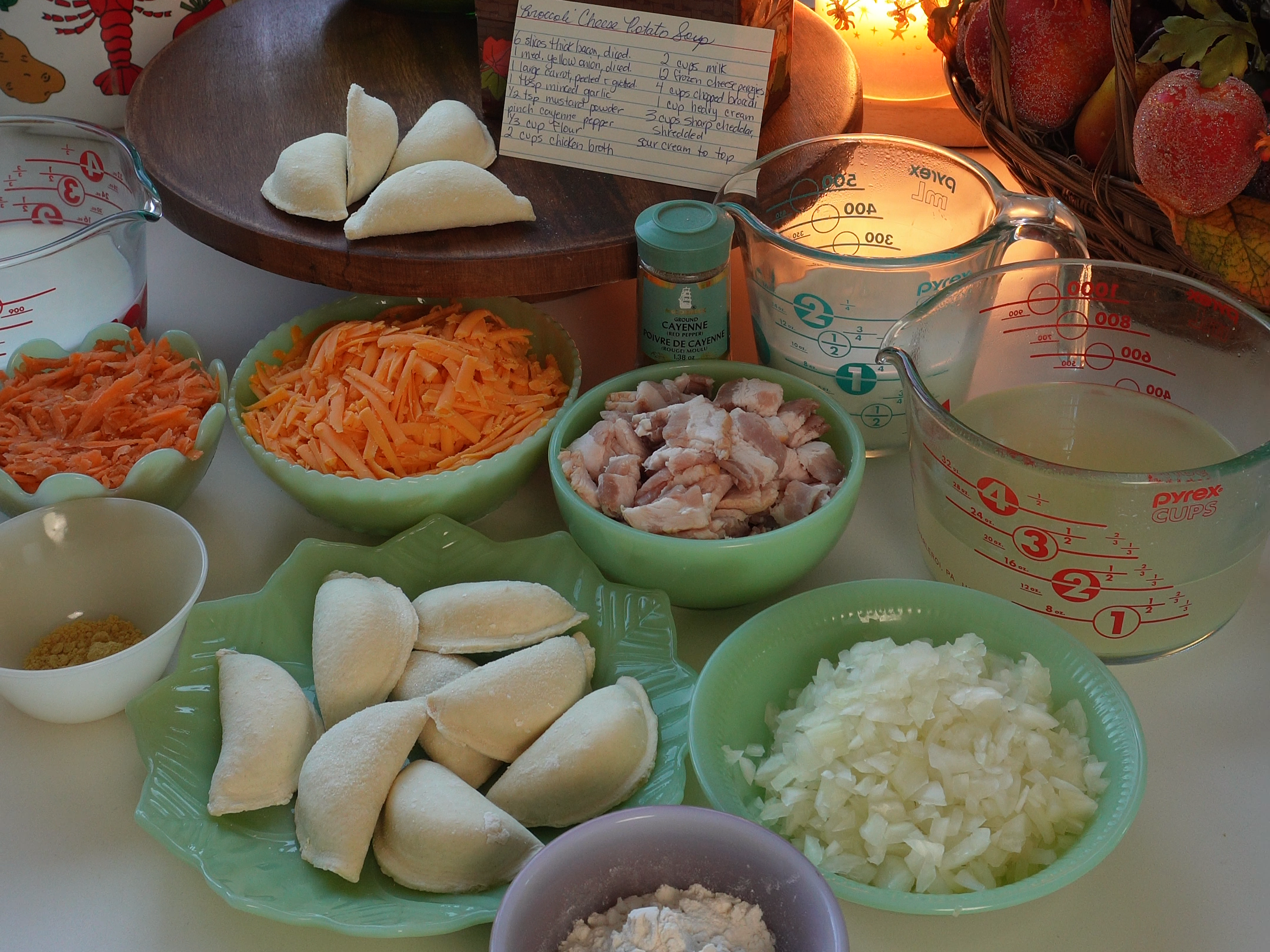 A kitchen table with chopped onions, cheese, flour, bacon, dough, and grated carrot on plates, surrounded by flowers, recipes, kitchen tools, candles, and measuring cups, lit by sunlight from a window.