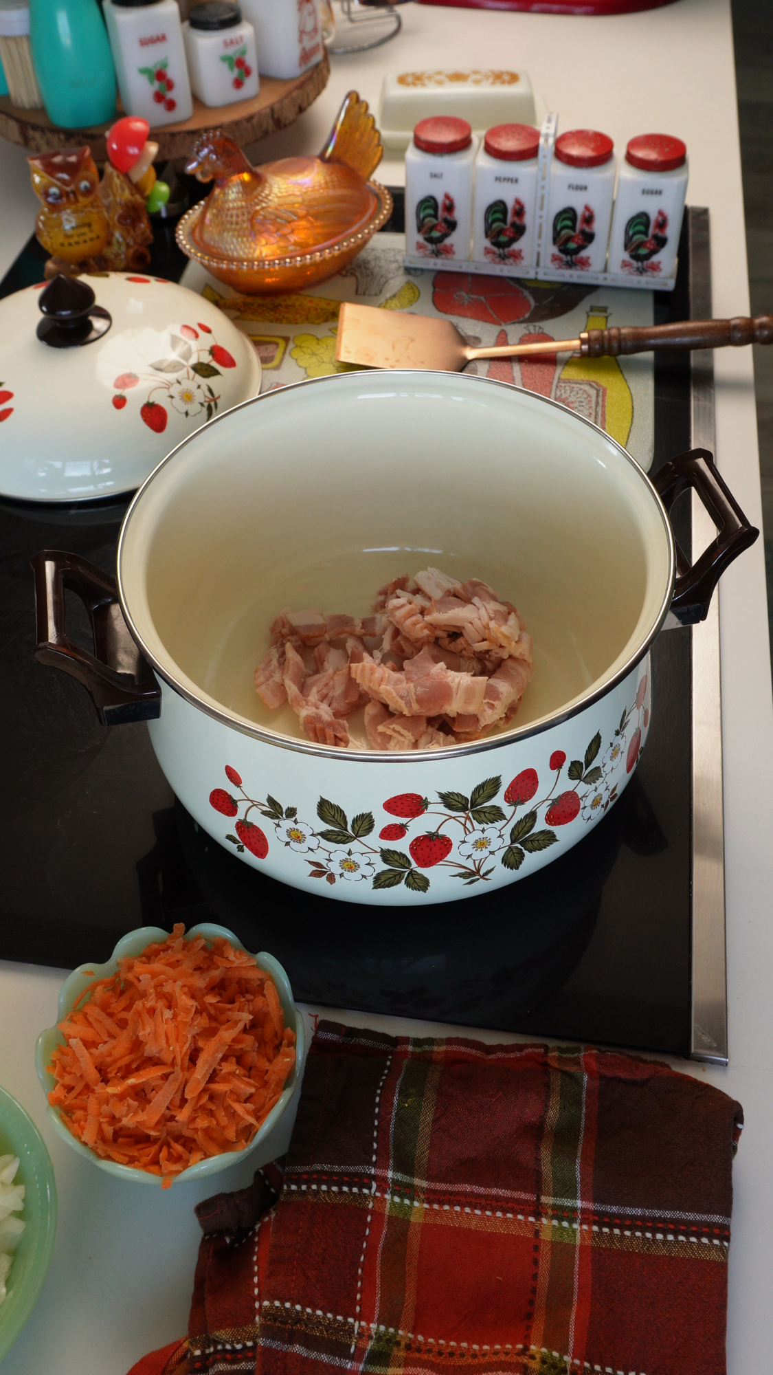 A large floral pot on a stove contains raw meat pieces. Chopped carrots and onions are nearby, with vintage kitchen items, towels, seasonings, and utensils around, creating a cozy and colorful cooking scene.