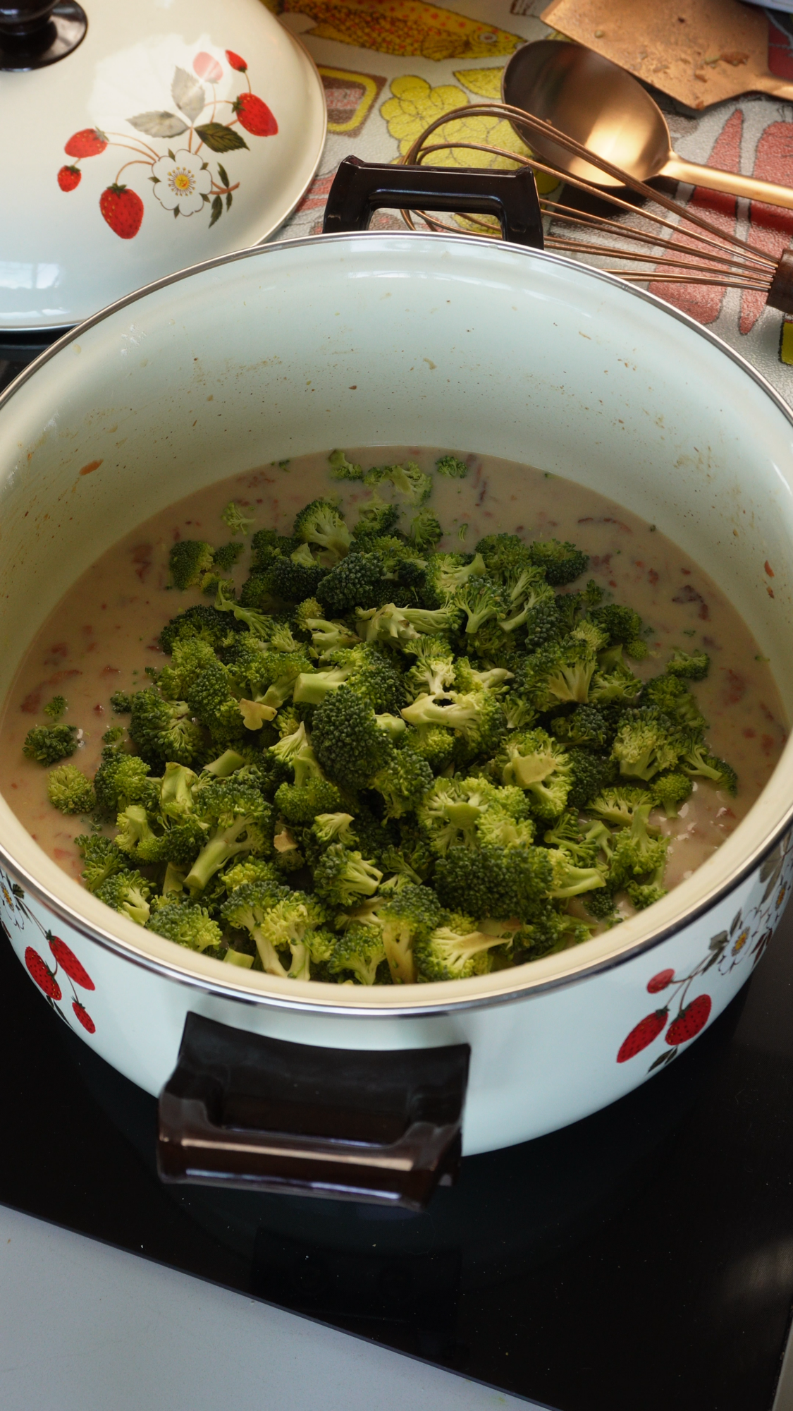 A white pot with a floral pattern contains a creamy mixture topped with fresh broccoli florets, sitting on a stovetop. The pot lid and kitchen utensils are nearby.