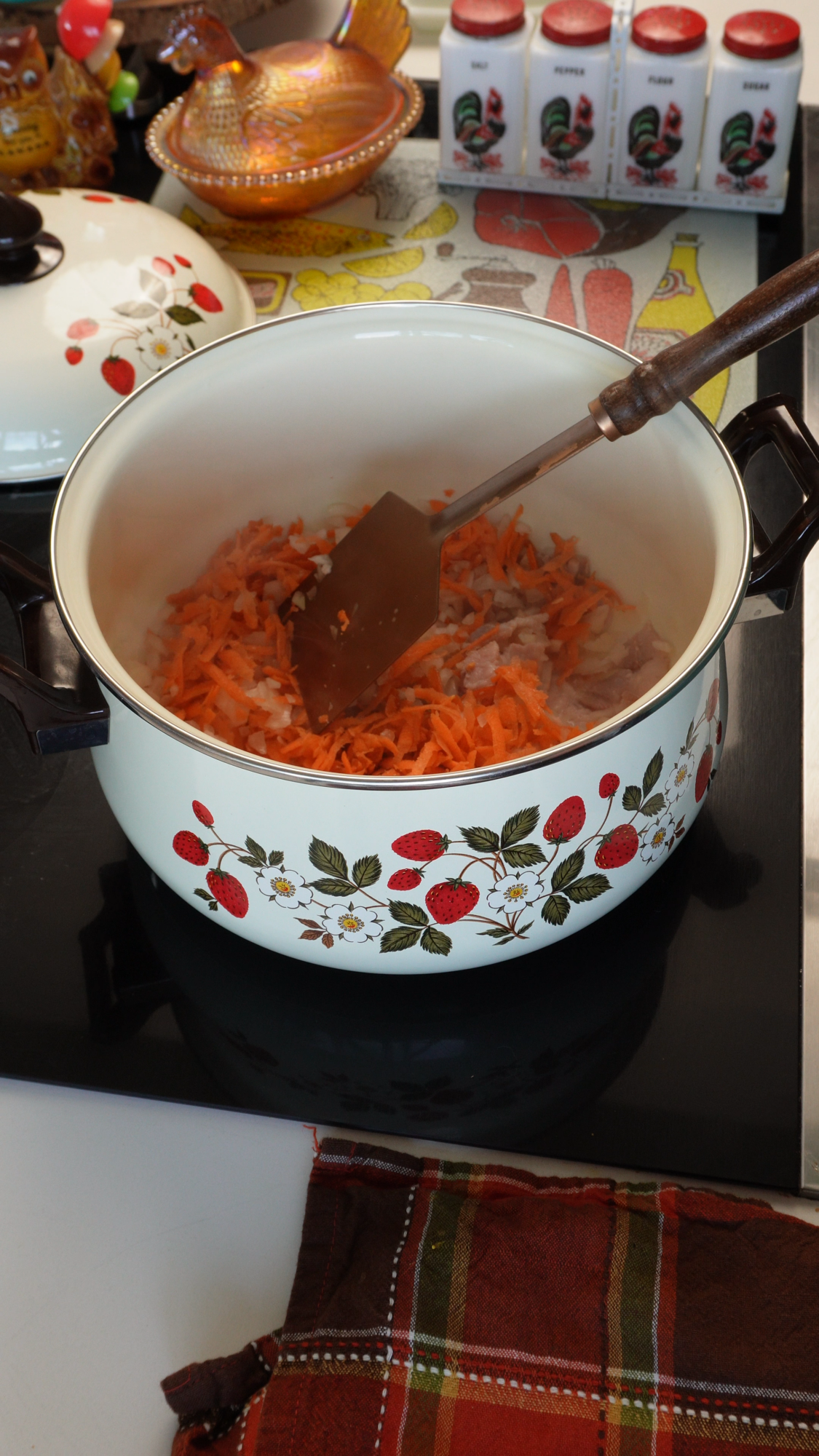 A white pot with a strawberry pattern contains chopped onions and grated carrots being stirred with a spatula. Nearby are spice jars, a plaid cloth, and a bowl on a kitchen counter.