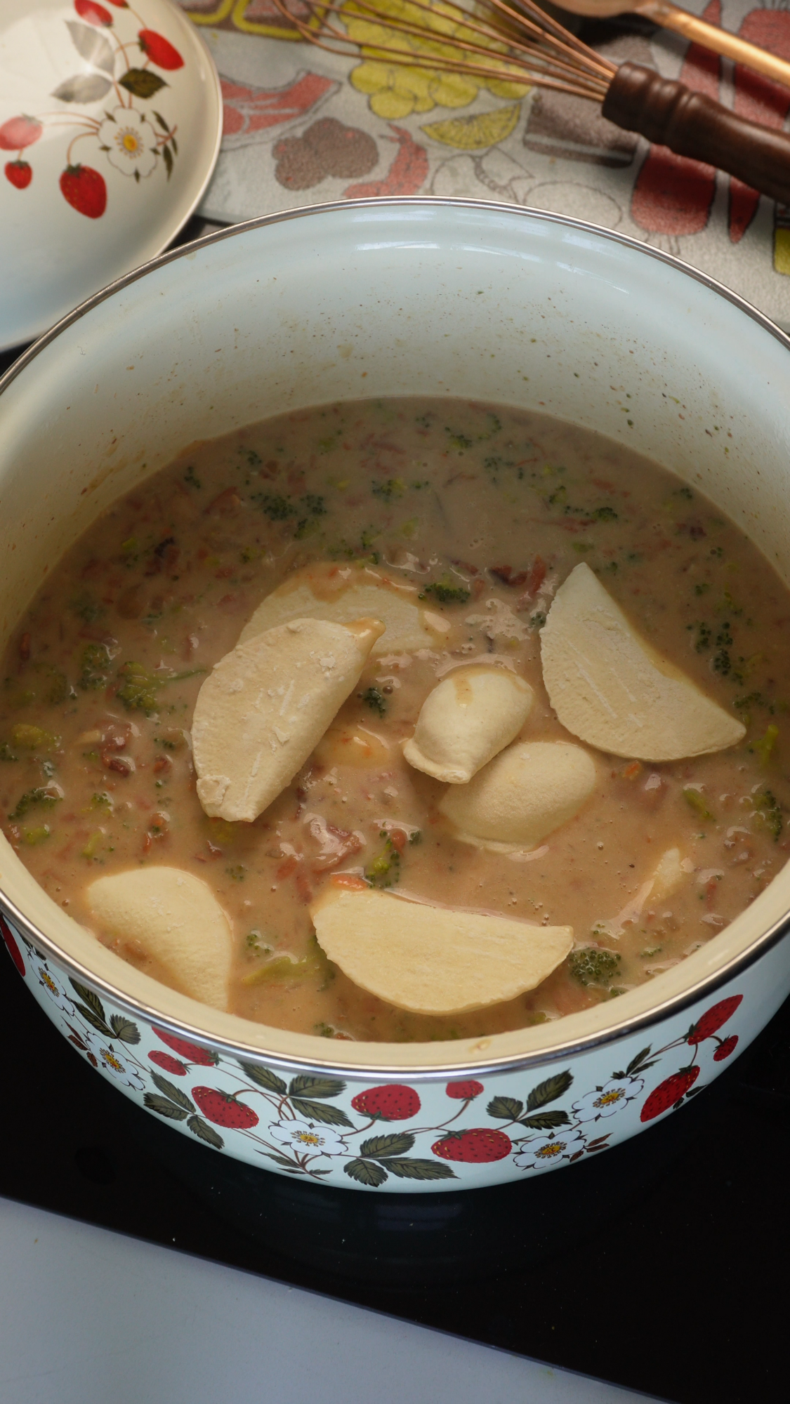 A pot of creamy soup with vegetables and several dumplings cooking inside, shown in a white pot decorated with red strawberries and green leaves.