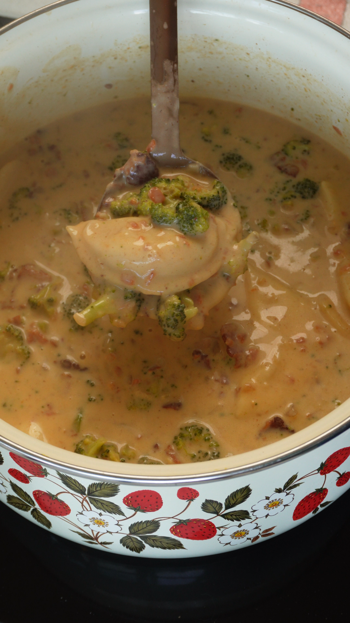 A ladle lifts creamy soup with broccoli and bacon from a white pot decorated with red strawberries and green leaves.