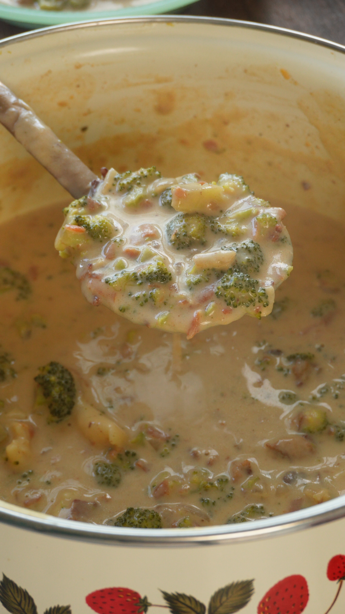 A ladle scoops creamy broccoli and cheese soup with chunks of broccoli and visible bits of bacon from a pot decorated with strawberries.