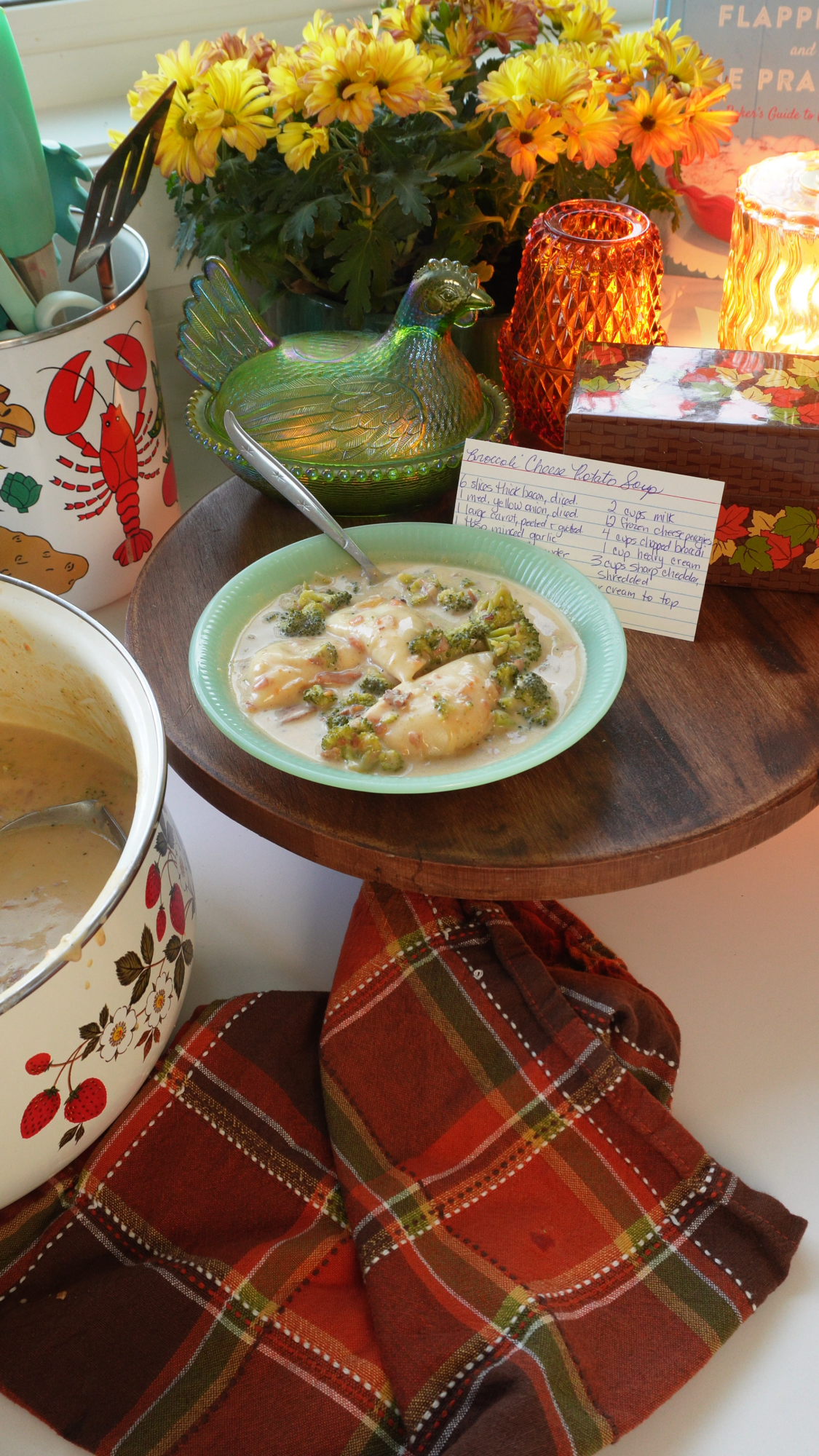 A bowl of creamy chicken and broccoli on a wooden board, with a recipe card, glass hen dish, plaid napkin, flowers, and vintage kitchenware nearby. Warm sunlight streams through the window.