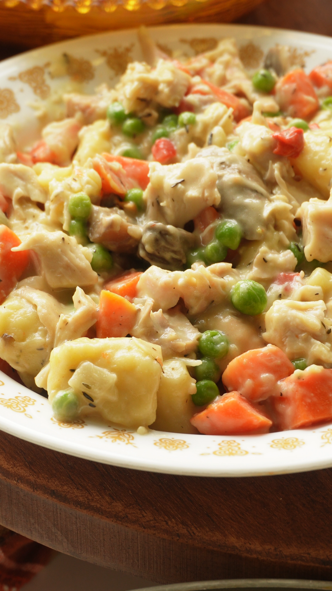 A close-up, top-down view of a creamy chicken pot pie filling featuring chunks of chicken, diced orange carrots, bright green peas, and potatoes in a savory sauce served on a white plate with gold decorative trim.