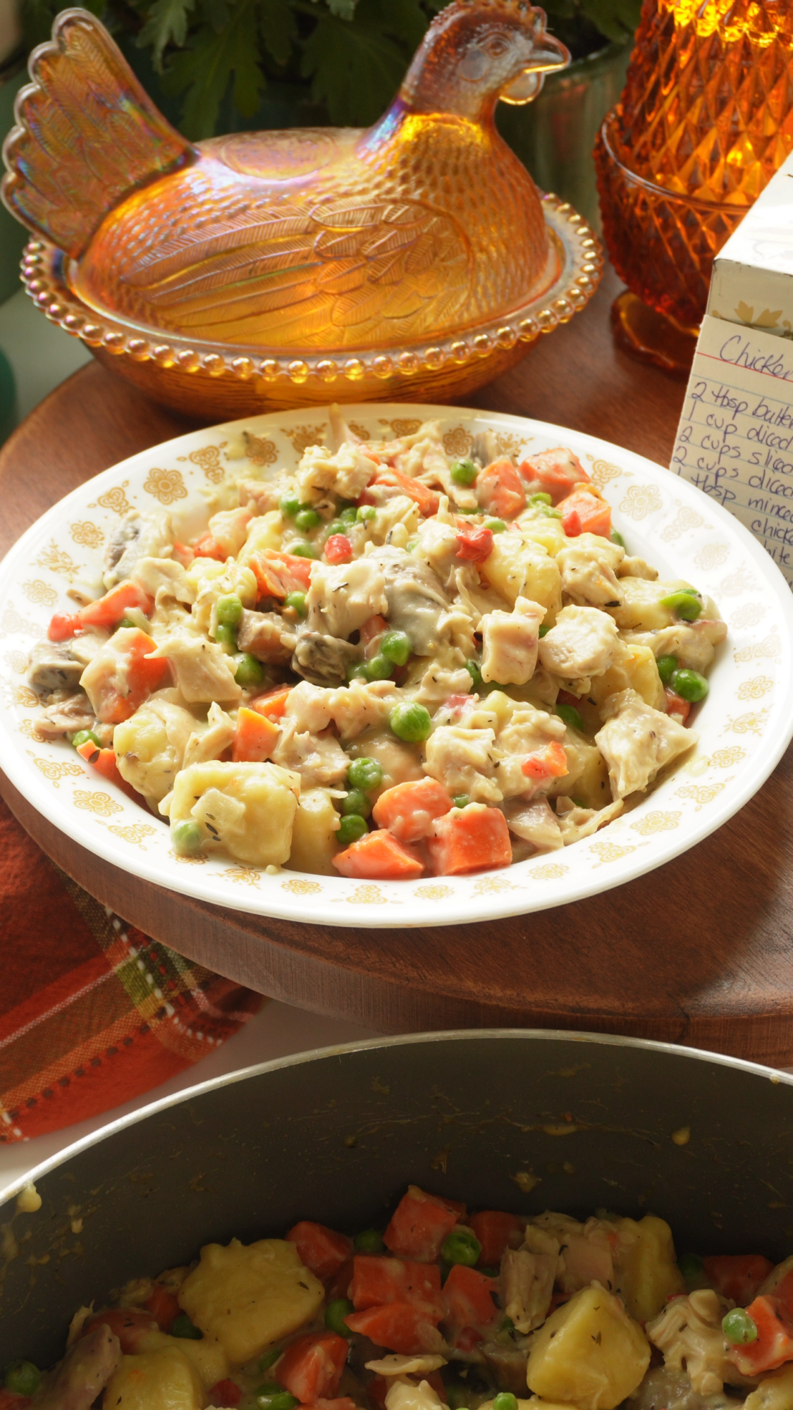 A plate of creamy chicken salad with potatoes, carrots, peas, and mushrooms sits on a wooden table next to a decorative glass dish and a handwritten recipe card.