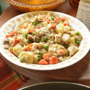 A plate of creamy chicken salad with potatoes, carrots, peas, and mushrooms sits on a wooden table next to a decorative glass dish and a handwritten recipe card.