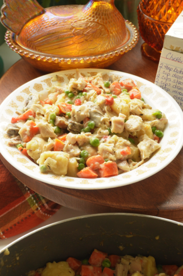 A plate of creamy chicken salad with potatoes, carrots, peas, and mushrooms sits on a wooden table next to a decorative glass dish and a handwritten recipe card.