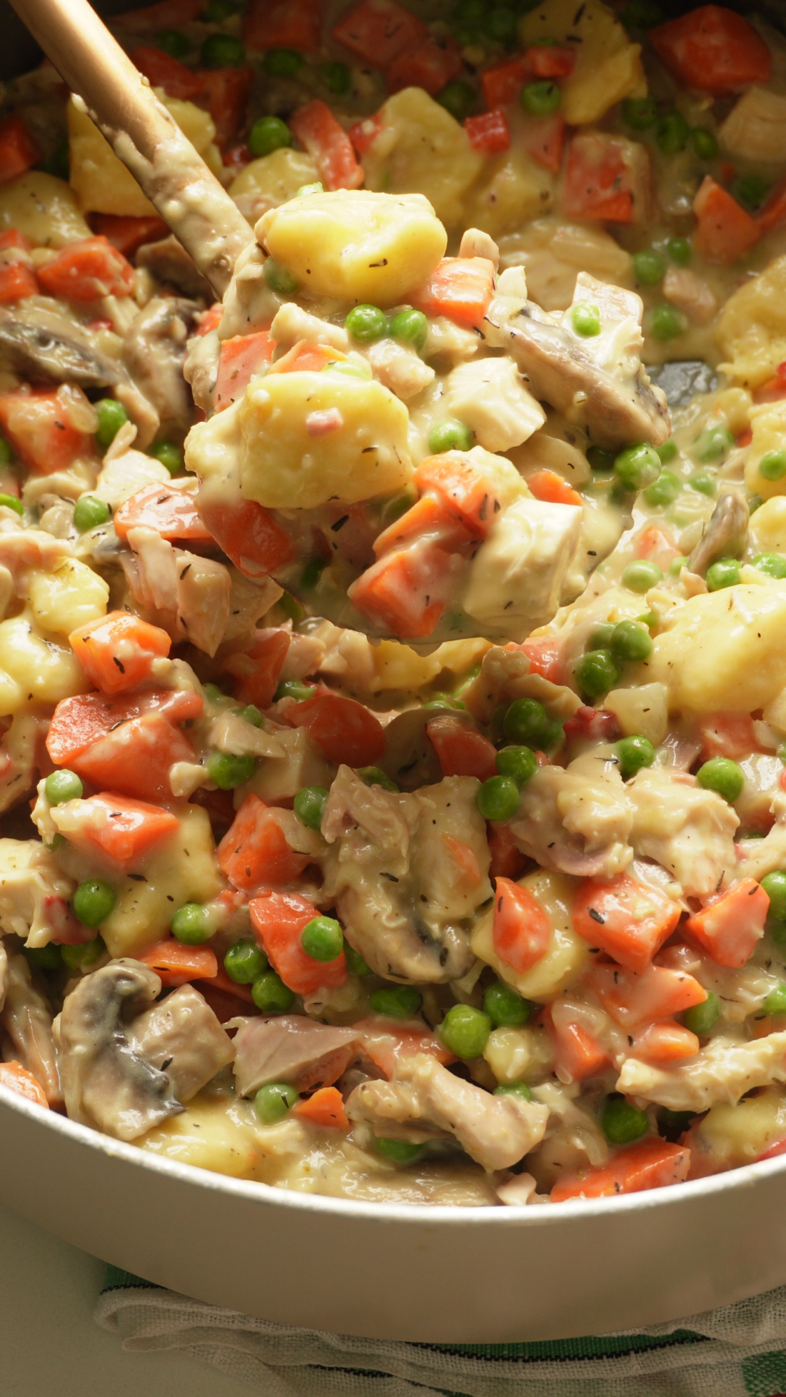 Close-up of a creamy mixed vegetable and chicken stew with peas, carrots, potatoes, and mushrooms being stirred with a wooden spoon.