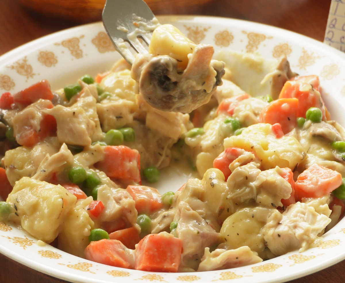 A close-up of a plate with creamy chicken and vegetable stew, featuring carrots, peas, mushrooms, and chunks of chicken. A fork is lifting a bite from the plate.