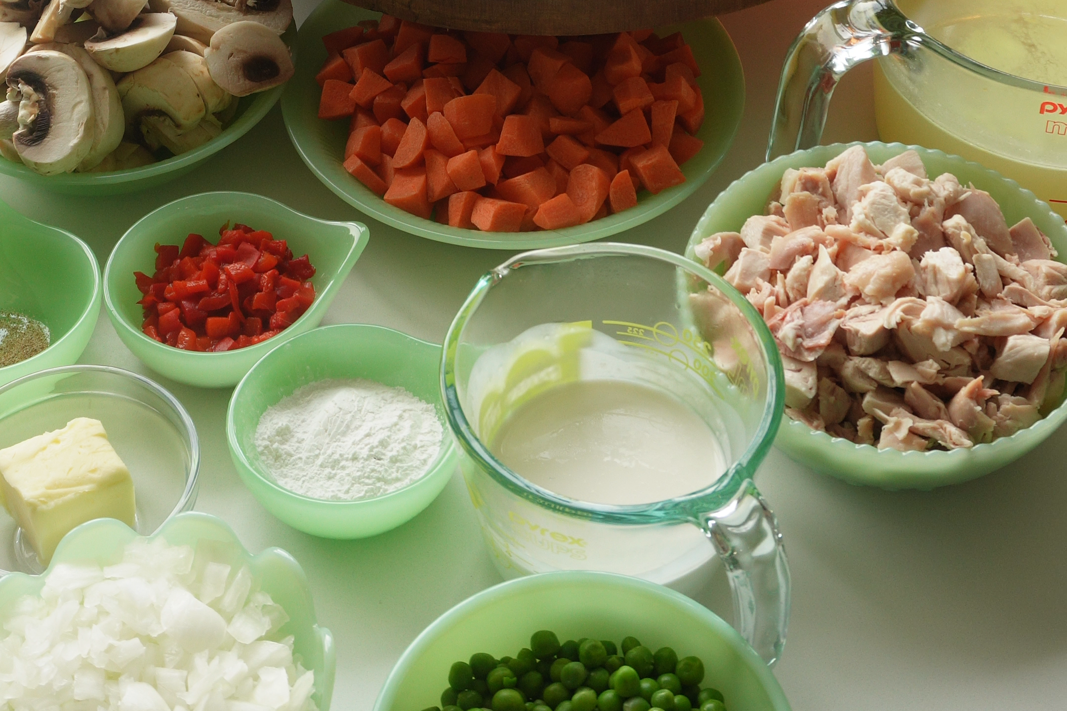 An assortment of prepped ingredients in green bowls, including chopped carrots, peas, mushrooms, bacon, butter, and milk, surround a wooden cake stand and amber glassware on a kitchen counter lit by candles.