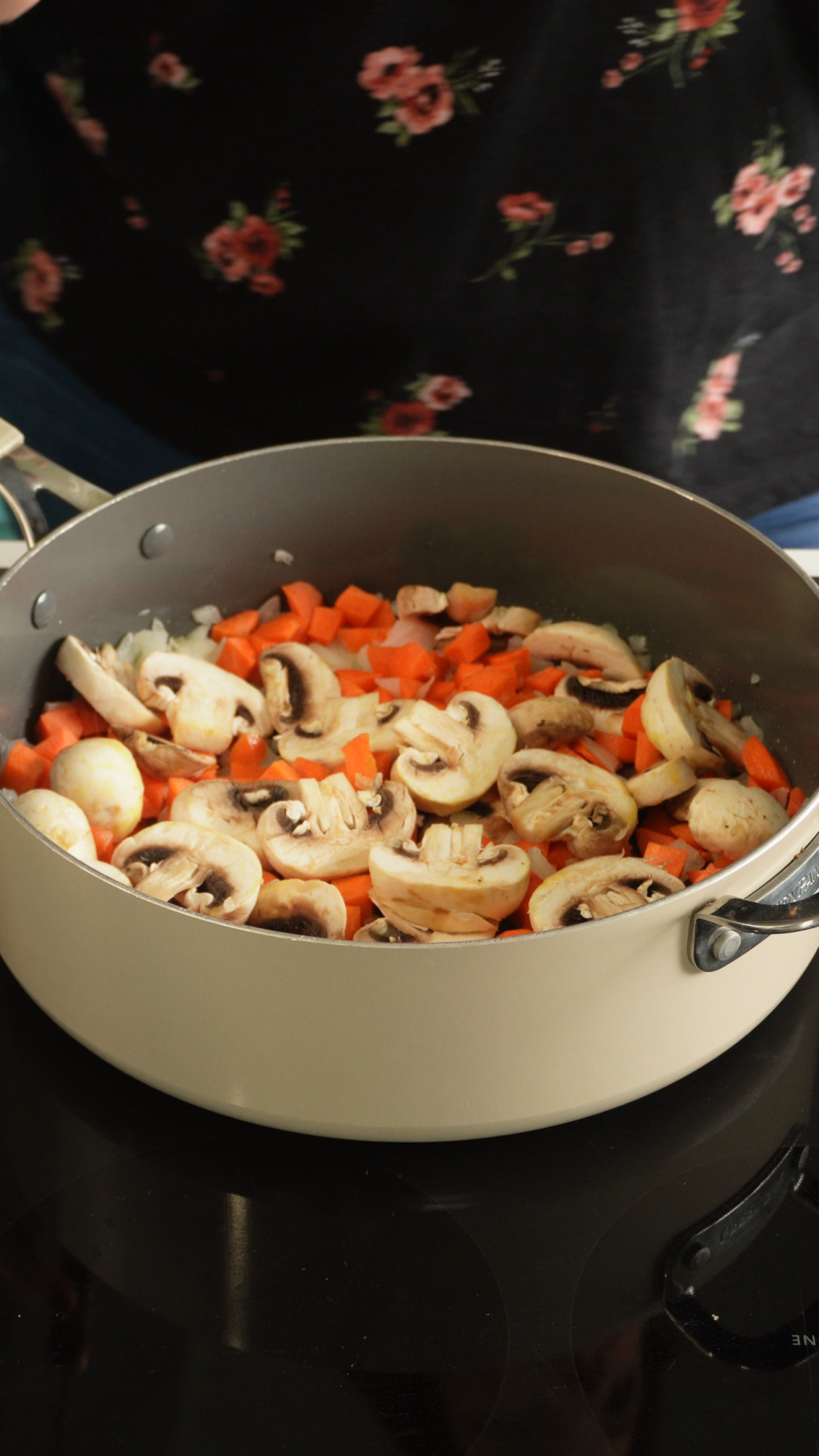 A saucepan on a stove contains sliced mushrooms, diced carrots, and chopped onions. A person in a floral shirt stands nearby, partially visible.