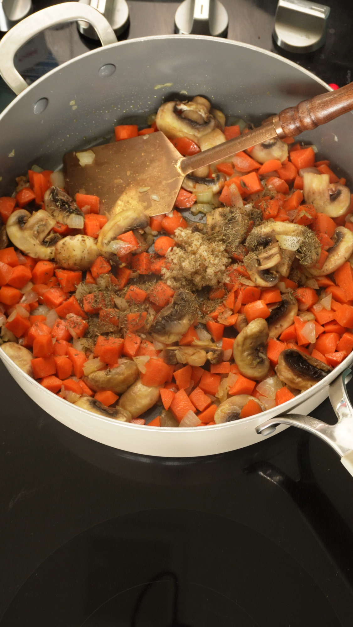 Diced carrots and sliced mushrooms being sautéed with spices in a large silver pan on a stovetop, stirred with a metal spatula.