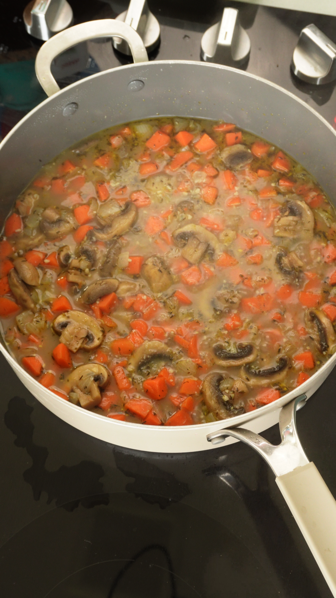 A white pan on a black stovetop contains a mixture of sliced mushrooms, diced carrots, and herbs simmering in a broth.