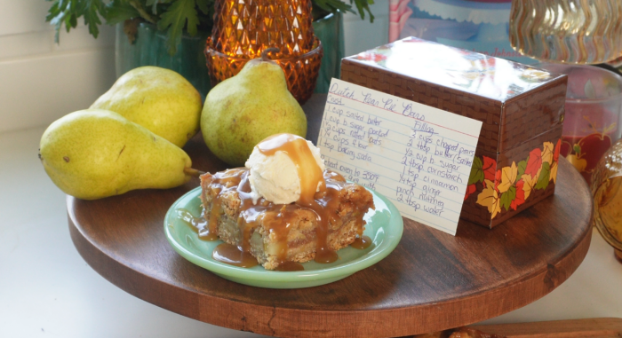 A dessert topped with ice cream and caramel sauce sits on a plate beside fresh pears, a handwritten recipe card, and a decorative box, all on a round wooden tray.