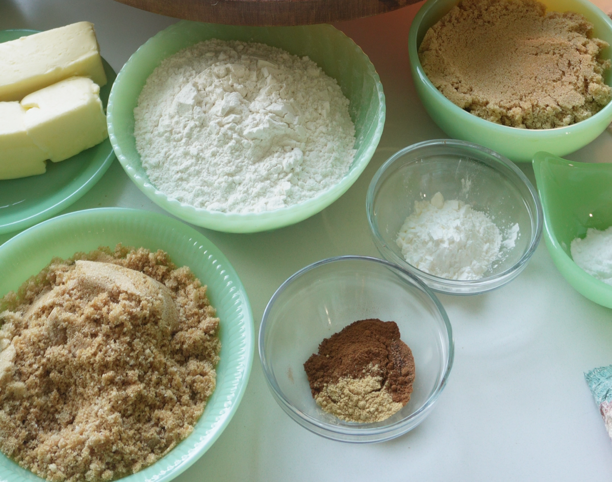 Bowls filled with flour, brown sugar, white sugar, butter, cornstarch, baking powder, and spices are arranged on a white countertop, ready to be used for baking.