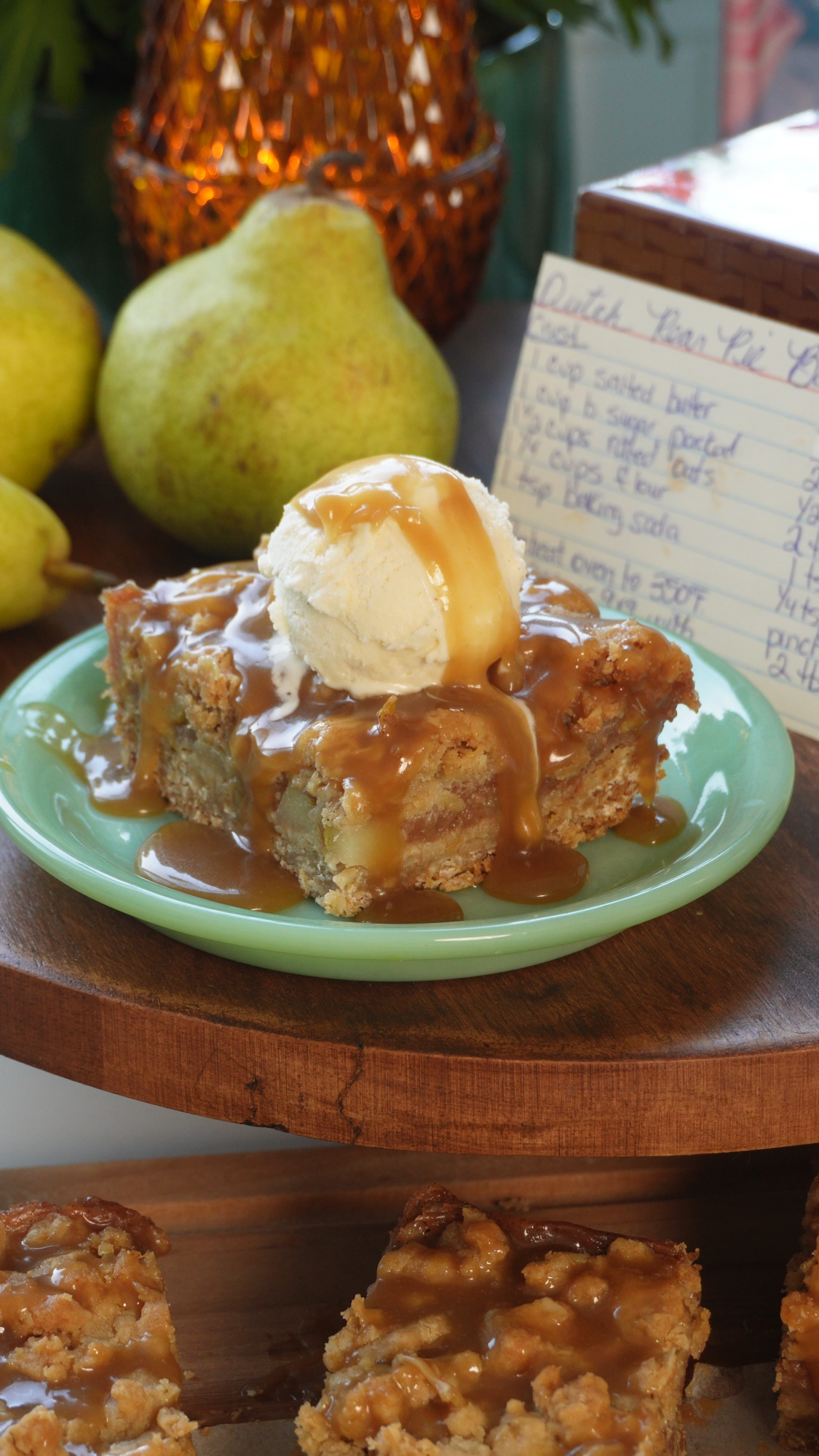 A slice of pear cake topped with a scoop of vanilla ice cream and caramel sauce sits on a green plate, with fresh pears and a handwritten recipe card in the background.