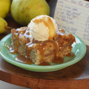 A slice of pear cake topped with a scoop of vanilla ice cream and caramel sauce sits on a green plate, with fresh pears and a handwritten recipe card in the background.
