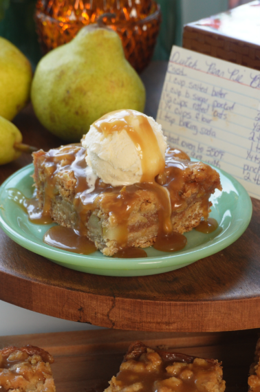 A slice of pear cake topped with a scoop of vanilla ice cream and caramel sauce sits on a green plate, with fresh pears and a handwritten recipe card in the background.