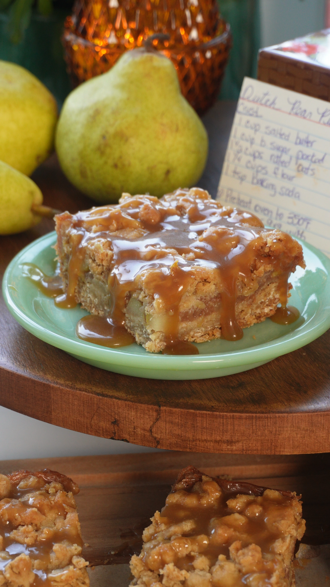 A slice of pear cake drizzled with caramel sauce sits on a green plate. Whole pears and a handwritten recipe card are in the background.