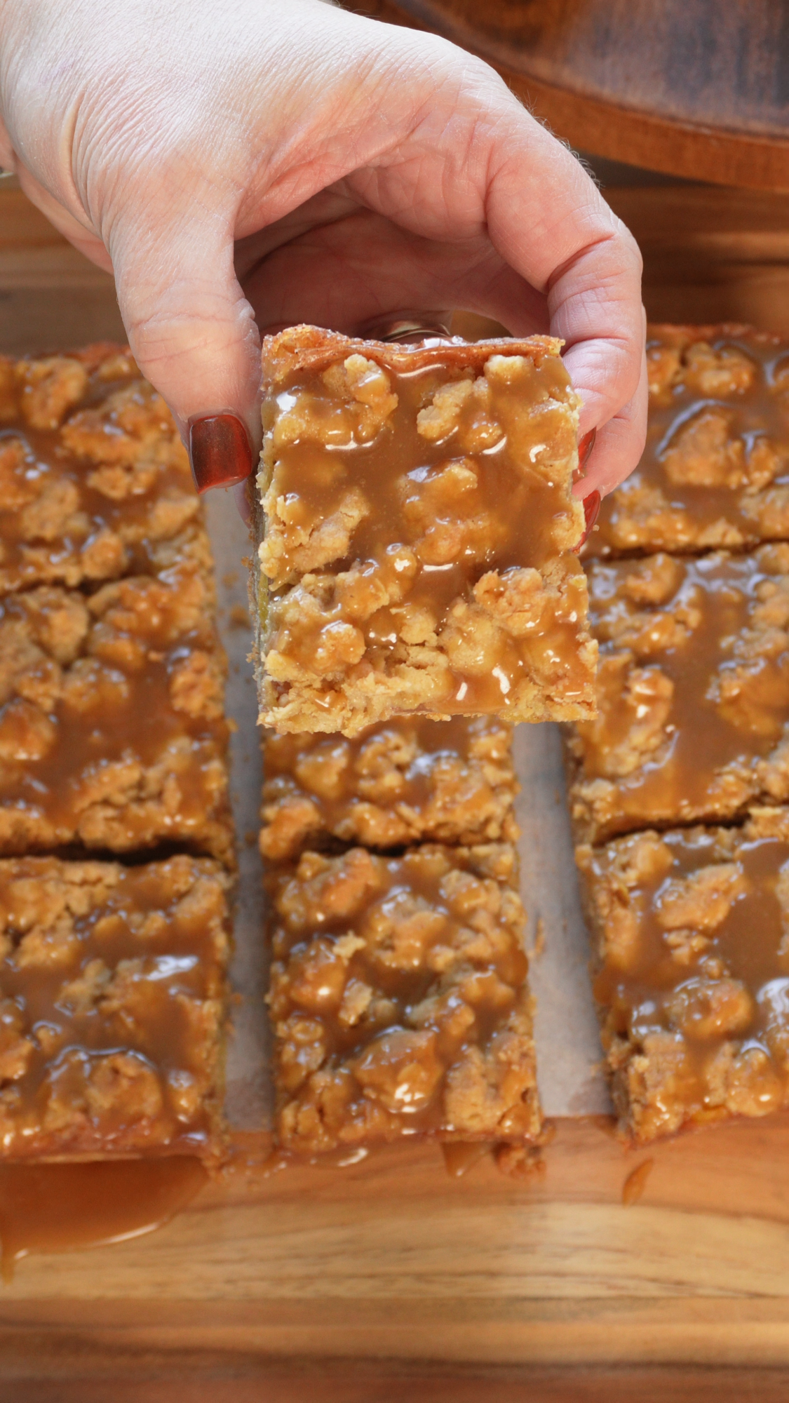 A hand with painted nails holds up a caramel-glazed oat bar above a wooden board with more oat bars, all topped with a shiny caramel layer.
