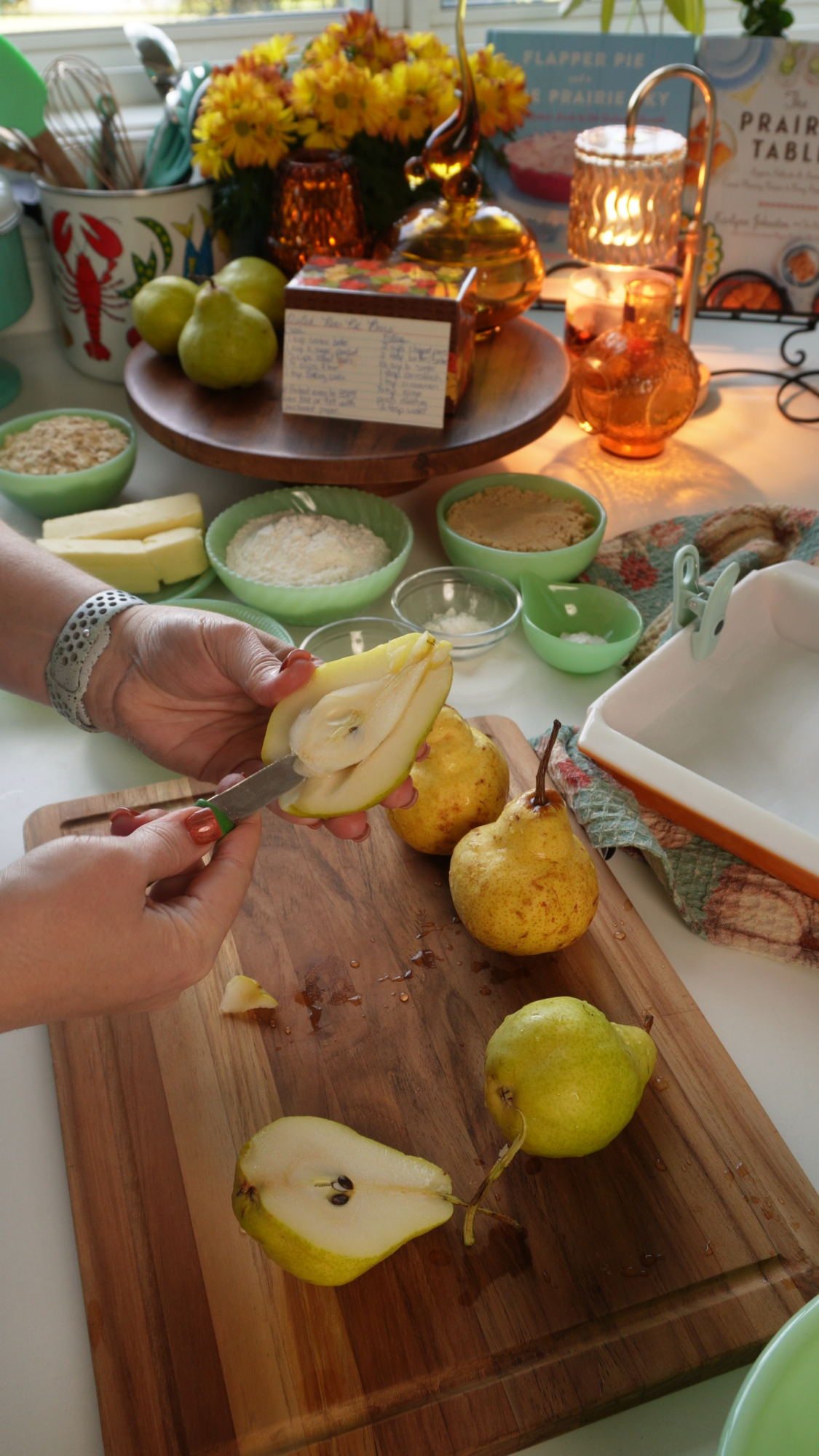 A person slices a pear on a wooden cutting board surrounded by pears, green bowls, a recipe card, flowers, and kitchen items on a table. Warm lighting creates a cozy atmosphere.